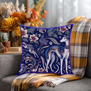 May include: A square throw pillow featuring a dark blue background with a repeating pattern of white and grey sighthounds, stylized floral motifs in shades of pink and blue, and small white paw prints. The pillow has a distinct blue piped edge.