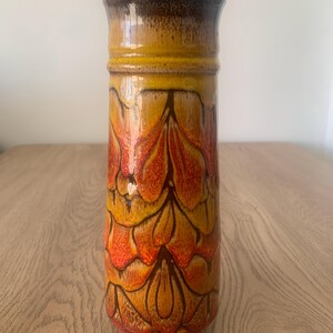 May include: A cylindrical ceramic vase featuring a glossy glaze with a repeating pattern of overlapping petal-like shapes in shades of orange, yellow, and brown. The top rim is finished in a dark brown tone, transitioning into a warm, vibrant gradient.
