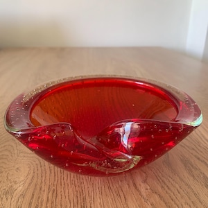 May include: A vibrant red glass bowl featuring a thick, clear glass rim and a distinctive bullicante technique with numerous small air bubbles suspended throughout the material. The bowl has a wide, shallow basin and a smooth, polished finish.