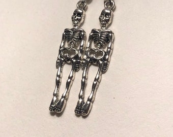 Puede incluir: Un par de pendientes de esqueleto plateados con un diseño de esqueleto colgante. Los pendientes tienen un cierre de gancho.