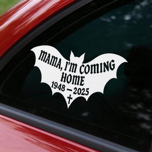 Puede incluir: Calcoman&iacute;a de vinilo blanco con forma de murci&eacute;lago, adherida a la ventana de un coche. La calcoman&iacute;a dice "MAMA, I'M COMING HOME" encima de "1948-2025" y una cruz. Fondo oscuro.