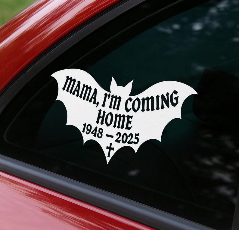 Puede incluir: Calcoman&iacute;a de vinilo blanco con forma de murci&eacute;lago, adherida a la ventana de un coche. La calcoman&iacute;a dice "MAMA, I'M COMING HOME" encima de "1948-2025" y una cruz. Fondo oscuro.