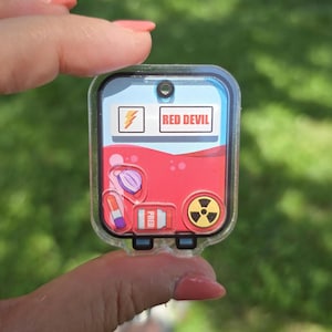 May include: A clear, rectangular keychain with a red liquid design and the text "RED DEVIL." The keychain features a lightning bolt symbol, a medical mask, a lipstick, and a radiation symbol. The keychain is held up against a blurred green background.