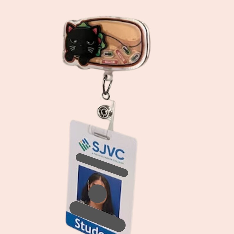以下が含まれることがあります： 黒猫とタコスデザインの格納式バッジリール。 「SJVC」と「Student」の文字が入った白いIDバッジに取り付けられています。 バッジには写真が表示されています。