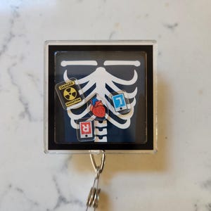 May include: A black and white x-ray image of a skeleton with a red heart, a blue phone, a yellow radiation symbol, and a red 'R' on a black background. The image is in a clear plastic frame with a silver retractable badge reel.