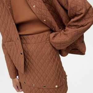 May include: A brown quilted jacket and skirt set. The jacket has a button closure and a patch pocket. The skirt has a high waist and a mini length. The set is made of a soft, quilted fabric.