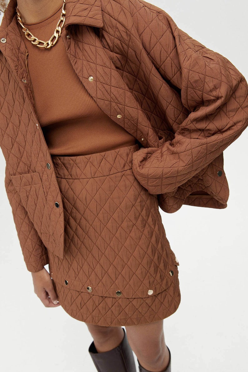 May include: A brown quilted jacket and skirt set. The jacket has a button closure and a patch pocket. The skirt has a high waist and a mini length. The set is made of a soft, quilted fabric.
