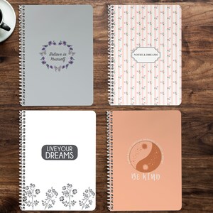 May include: Four spiral bound notebooks with different designs. The first notebook is gray with a floral wreath and the text "Believe in Yourself". The second notebook has a pink and white striped pattern with small pink flowers and the text "Notes & Dreams". The third notebook is white with a gray floral border and the text "Live Your Dreams". The fourth notebook is peach with a brown and white yin yang symbol and the text "Be Kind".