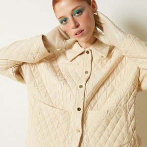 May include: A cream-colored quilted jacket with a button-up front and a patch pocket. The jacket is made of a soft, textured fabric and has a relaxed fit.