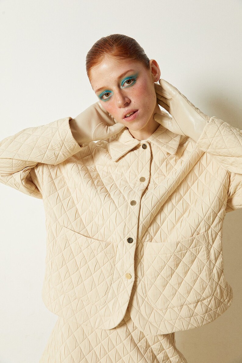 May include: A cream-colored quilted jacket with a button-up front and a patch pocket. The jacket is made of a soft, textured fabric and has a relaxed fit.