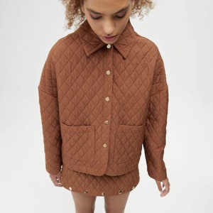 May include: A brown quilted jacket with a button-up front and a patch pocket on the chest. The jacket is paired with a matching brown quilted skirt with gold button accents.