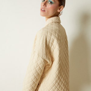 May include: A cream-colored quilted jacket with a high collar and long sleeves. The jacket is made of a soft, textured fabric and has a relaxed fit.