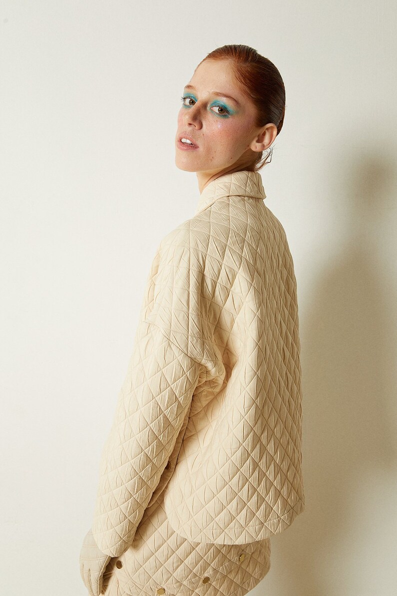 May include: A cream-colored quilted jacket with a high collar and long sleeves. The jacket is made of a soft, textured fabric and has a relaxed fit.