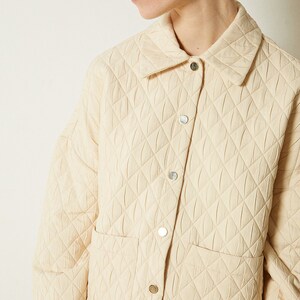 May include: A cream-colored quilted jacket with a button-up front and a patch pocket on the left side. The jacket has a diamond-shaped quilting pattern.