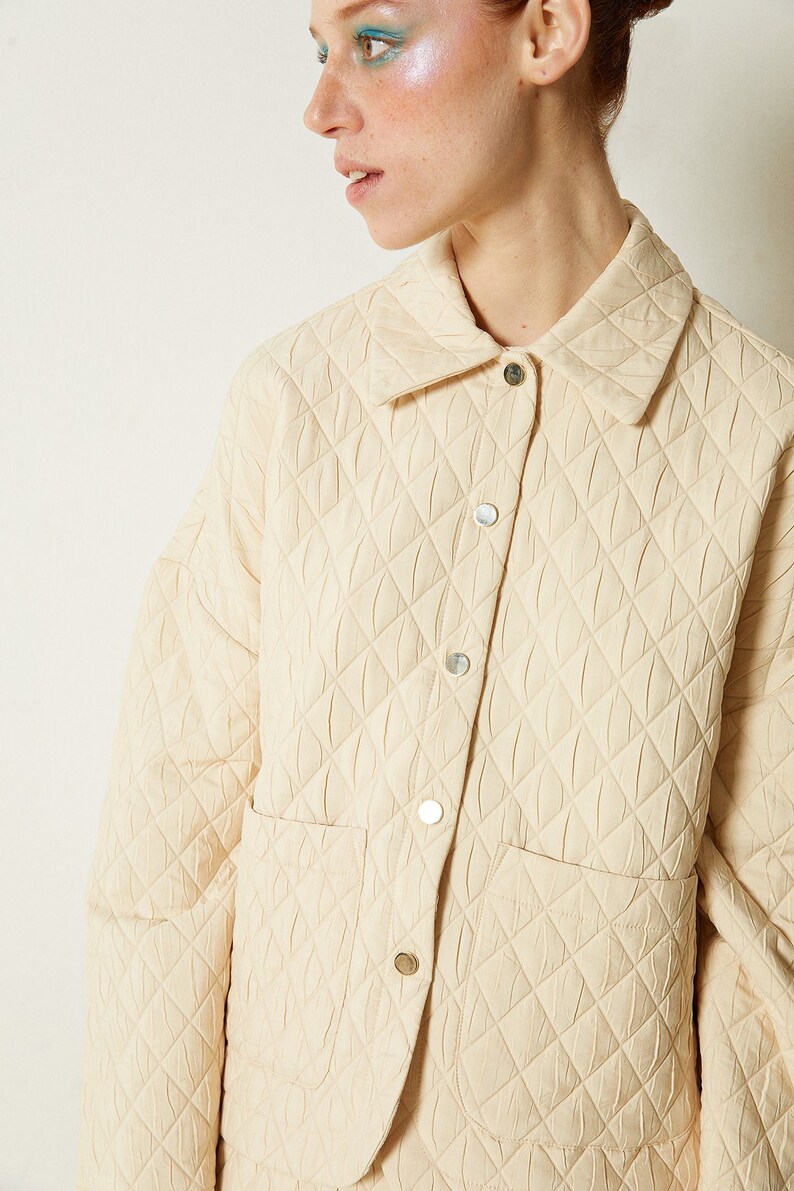 May include: A cream-colored quilted jacket with a button-up front and a patch pocket on the left side. The jacket has a diamond-shaped quilting pattern.