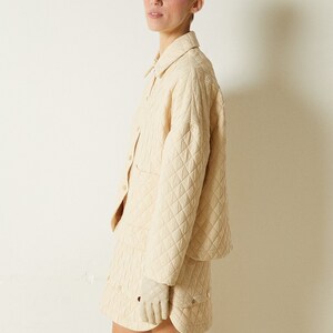 May include: A cream-colored quilted jacket and skirt set. The jacket has a button-up front and long sleeves. The skirt is a mini skirt with a slightly flared hem.
