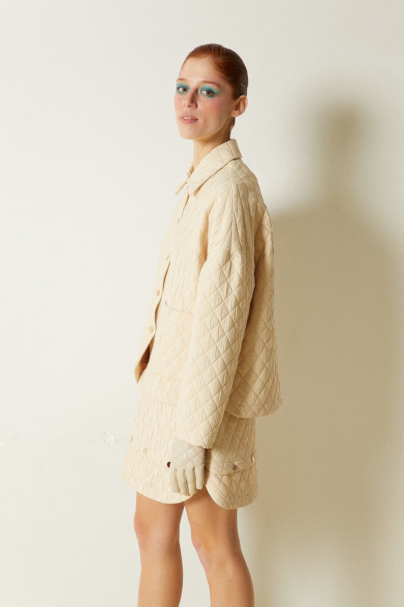 May include: A cream-colored quilted jacket and skirt set. The jacket has a button-up front and long sleeves. The skirt is a mini skirt with a slightly flared hem.