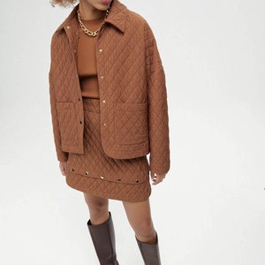May include: A brown quilted jacket with a snap closure and two front pockets. The jacket is paired with a brown quilted skirt with a studded trim. The model is wearing brown knee-high boots.