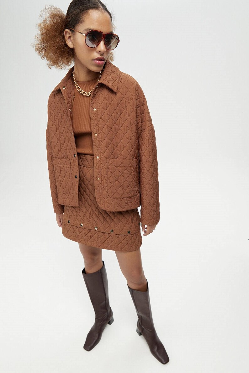 May include: A brown quilted jacket with a snap closure and two front pockets. The jacket is paired with a brown quilted skirt with a studded trim. The model is wearing brown knee-high boots.