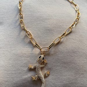 May include: Gold-toned chain necklace with a large link design. The pendant is a rhinestone-encrusted lizard with black eyes and gold-toned feet. The necklace is displayed on a neutral-colored surface.