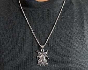 May include: A silver-toned metal pendant necklace featuring a detailed relief of a bearded figure wearing a horned helmet, flanked by two crossed swords. The pendant hangs from a thin, textured silver-toned chain against a black ribbed fabric.