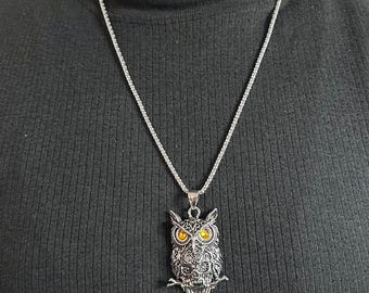 May include: A silver-toned metal necklace featuring an owl-shaped pendant with intricate filigree detailing and two prominent amber-colored circular eyes. The pendant hangs from a thin, polished silver-toned chain that rests against a black fabric.