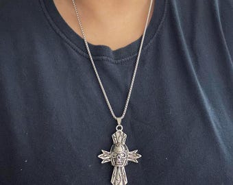 May include: A silver-toned metal pendant necklace featuring a cross design with an integrated skull at the center. The cross has textured, flame-like detailing on each arm and is suspended from a thin, silver-toned box chain necklace.