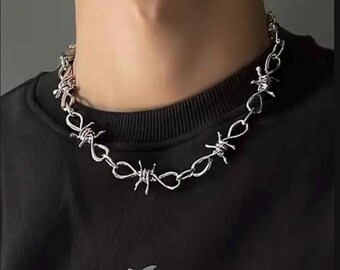May include: A silver-toned metal chain necklace featuring a repeating design of interconnected links shaped like barbed wire. The necklace rests against a black shirt, showcasing a polished, reflective finish on each individual wire segment.