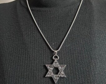 May include: A silver-toned Star of David pendant necklace featuring an intricate, engraved pattern on the six-pointed star. The pendant hangs from a thin, textured silver-colored chain, resting against a plain black ribbed fabric background.