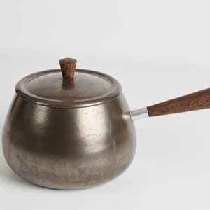 Puede incluir: Una olla de fondue de plata con un mango y una tapa de madera. La olla tiene una forma redondeada y una superficie lisa.