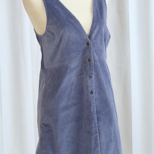 Puede incluir: Vestido de pana azul bígaro con cuello en V y botones delanteros. El vestido sin mangas se exhibe en un maniquí. La tela tiene estrías verticales.