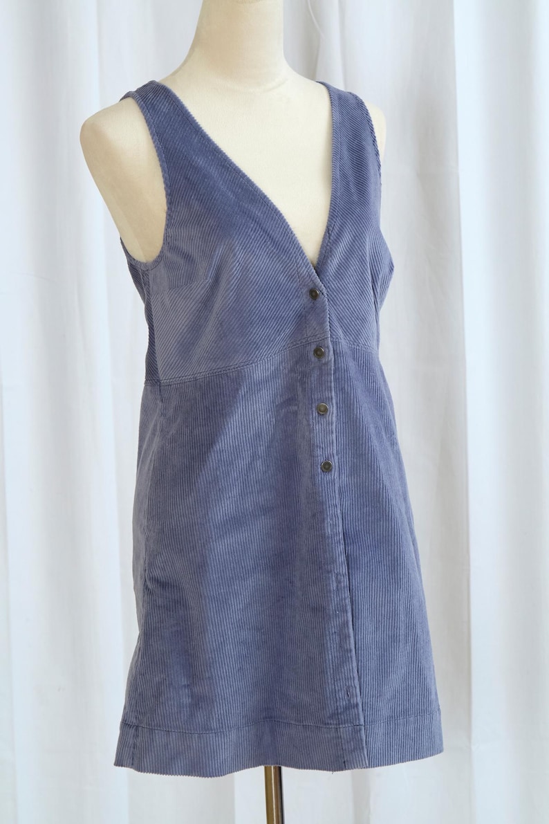 May include: A periwinkle blue corduroy dress with a V-neck and button-down front. The sleeveless dress is displayed on a mannequin. The fabric has vertical ridges.