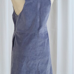 May include: A periwinkle blue corduroy overall dress. The dress features a scoop neckline, a sleeveless design with cut-out details, and a straight skirt. The fabric has vertical ridges.