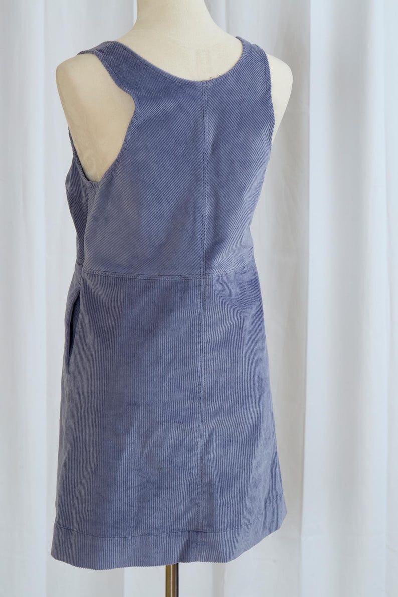 May include: A periwinkle blue corduroy overall dress. The dress features a scoop neckline, a sleeveless design with cut-out details, and a straight skirt. The fabric has vertical ridges.