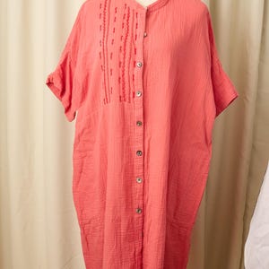 Puede incluir: Vestido color coral con mangas cortas y cuello mandarín. El vestido presenta una botonadura frontal y bordados decorativos en la parte superior del pecho. La tela tiene una apariencia texturizada, exhibido en un maniquí.