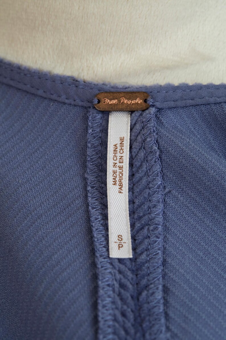 May include: Close-up of a blue garment with a textured fabric. A brown tag with the words "Free People" is at the top. A white tag with the text "MADE IN CHINA FABRIQU&Eacute; EN CHINE" and the size "S/P" is sewn into the garment.