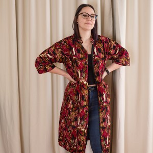 May include: A woman wearing a long, red, black and gold patterned kimono-style robe with a black top and blue jeans.