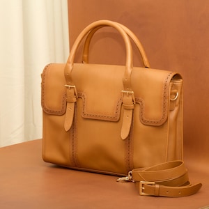 May include: A tan leather briefcase with two buckles and a strap. The briefcase has a stitched design and a flap closure.