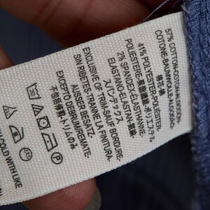 May include: A close-up of a white clothing tag with text in multiple languages, including English, French, and Japanese. The tag lists the fabric composition as cotton, polyester, and spandex. Washing instructions are also visible.