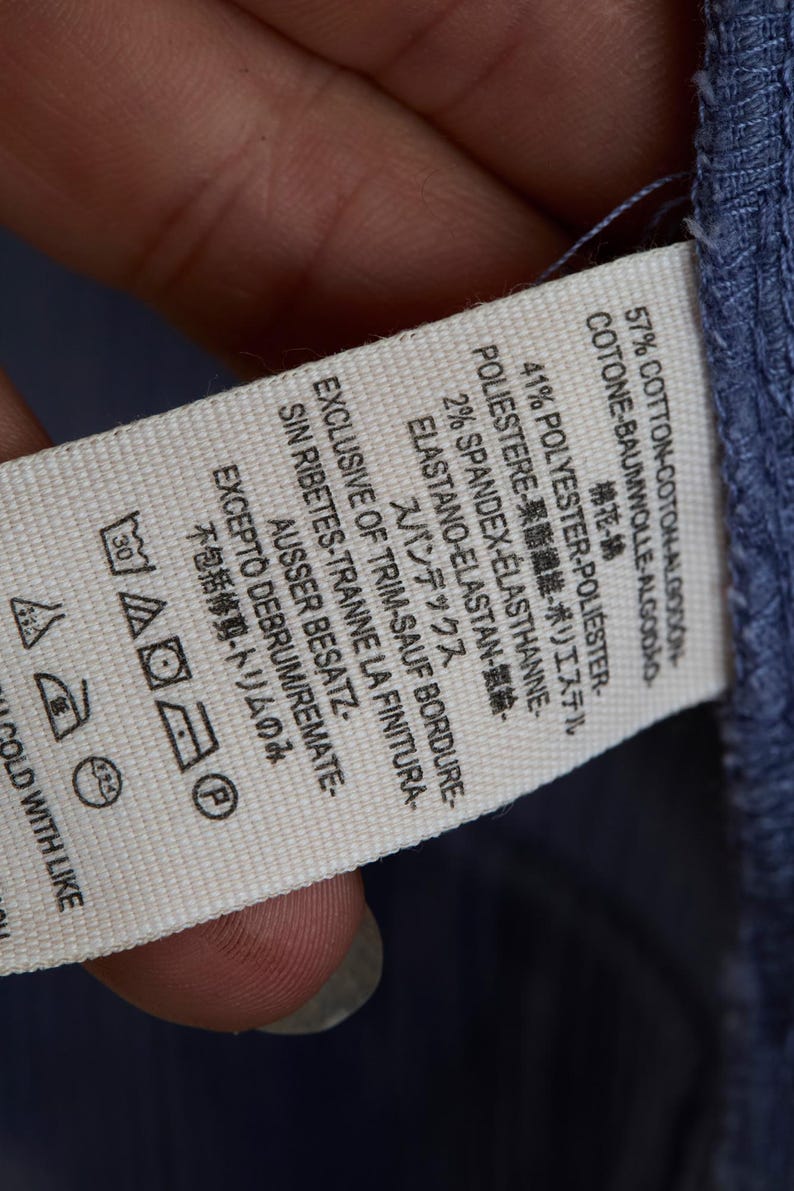 May include: A close-up of a white clothing tag with text in multiple languages, including English, French, and Japanese. The tag lists the fabric composition as cotton, polyester, and spandex. Washing instructions are also visible.