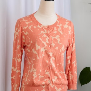 Op de afbeelding: Koraalkleurig vest met een crèmekleurig bloemmotief. De trui met lange mouwen heeft een ronde hals en is aan de voorkant dichtgeknoopt. Het vest wordt op een mannequin getoond.
