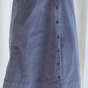 May include: A periwinkle blue corduroy dress with a button-up front. The dress has a sleeveless design and a subtle vertical ribbed texture. The buttons are a dark, muted color, and the dress appears to be knee-length.