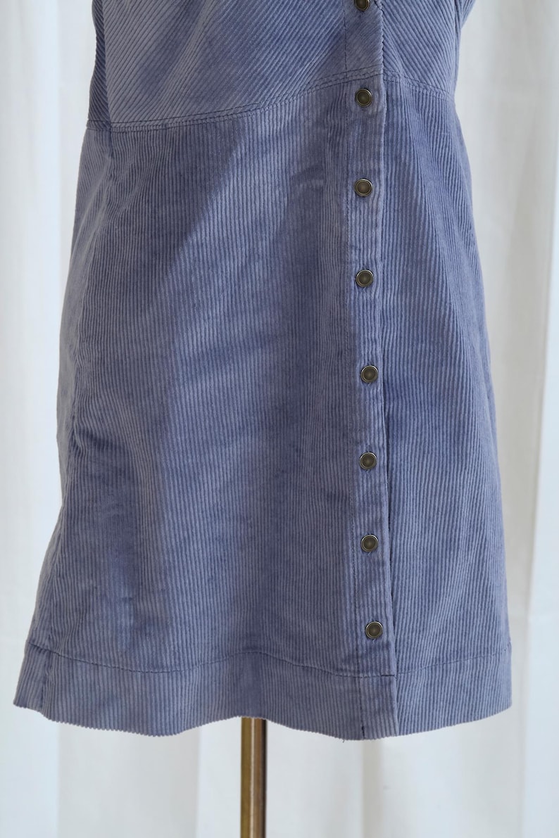 May include: A periwinkle blue corduroy dress with a button-up front. The dress has a sleeveless design and a subtle vertical ribbed texture. The buttons are a dark, muted color, and the dress appears to be knee-length.