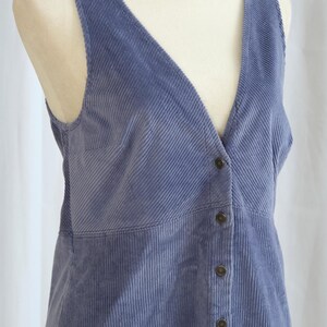May include: A periwinkle blue corduroy vest with a V-neck and button-down front. The vest is displayed on a mannequin and features a vertical ribbed texture. The vest has a tailored fit and a classic design.