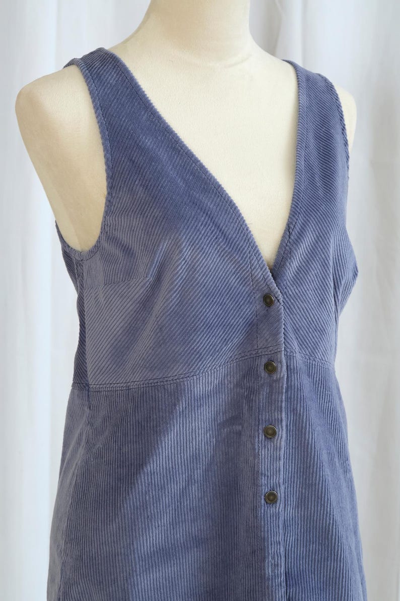 May include: A periwinkle blue corduroy vest with a V-neck and button-down front. The vest is displayed on a mannequin and features a vertical ribbed texture. The vest has a tailored fit and a classic design.
