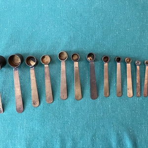 May include: A collection of antique brass measuring spoons of varying sizes, arranged in a row. Each spoon has a long, flat handle and a rounded bowl. The spoons are numbered from 1 to 8, with the largest spoon on the left. The spoons are on a teal surface.