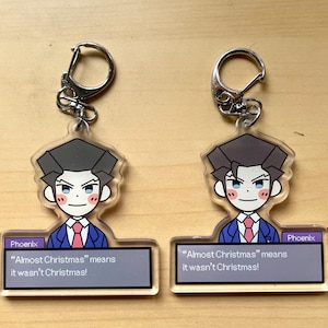 May include: Two clear acrylic keychains featuring a cartoon character with black hair, a blue suit, and a pink tie. The text "Phoenix" and "Almost Christmas" means it wasn't Christmas!" is printed on the keychain.