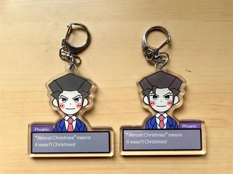 May include: Two clear acrylic keychains featuring a cartoon character with black hair, a blue suit, and a pink tie. The text "Phoenix" and "Almost Christmas" means it wasn't Christmas!" is printed on the keychain.