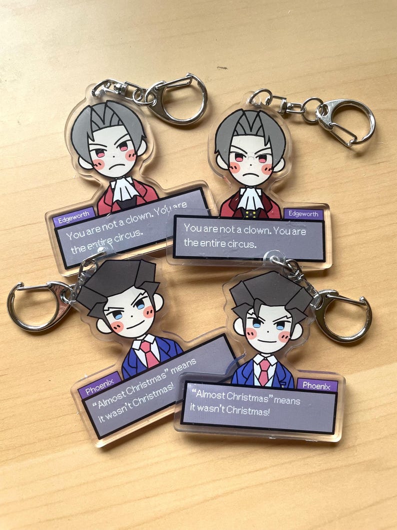May include: Four clear acrylic keychains featuring characters from the Ace Attorney video game series. Each keychain has a different character and a quote from the game. The characters are Miles Edgeworth, Phoenix Wright, and two more Miles Edgeworth keychains. The keychains are on a wooden surface.