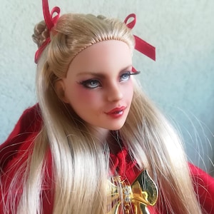 Könnte beinhalten: Eine Puppe mit langem blondem Haar, roten Schleifen und einem roten Kapuzenmantel. Die Puppe hat blaue Augen, rote Lippen und einen goldenen Schleifenanhänger. Das Haar der Puppe ist mit einem Seitenscheitel gestylt.